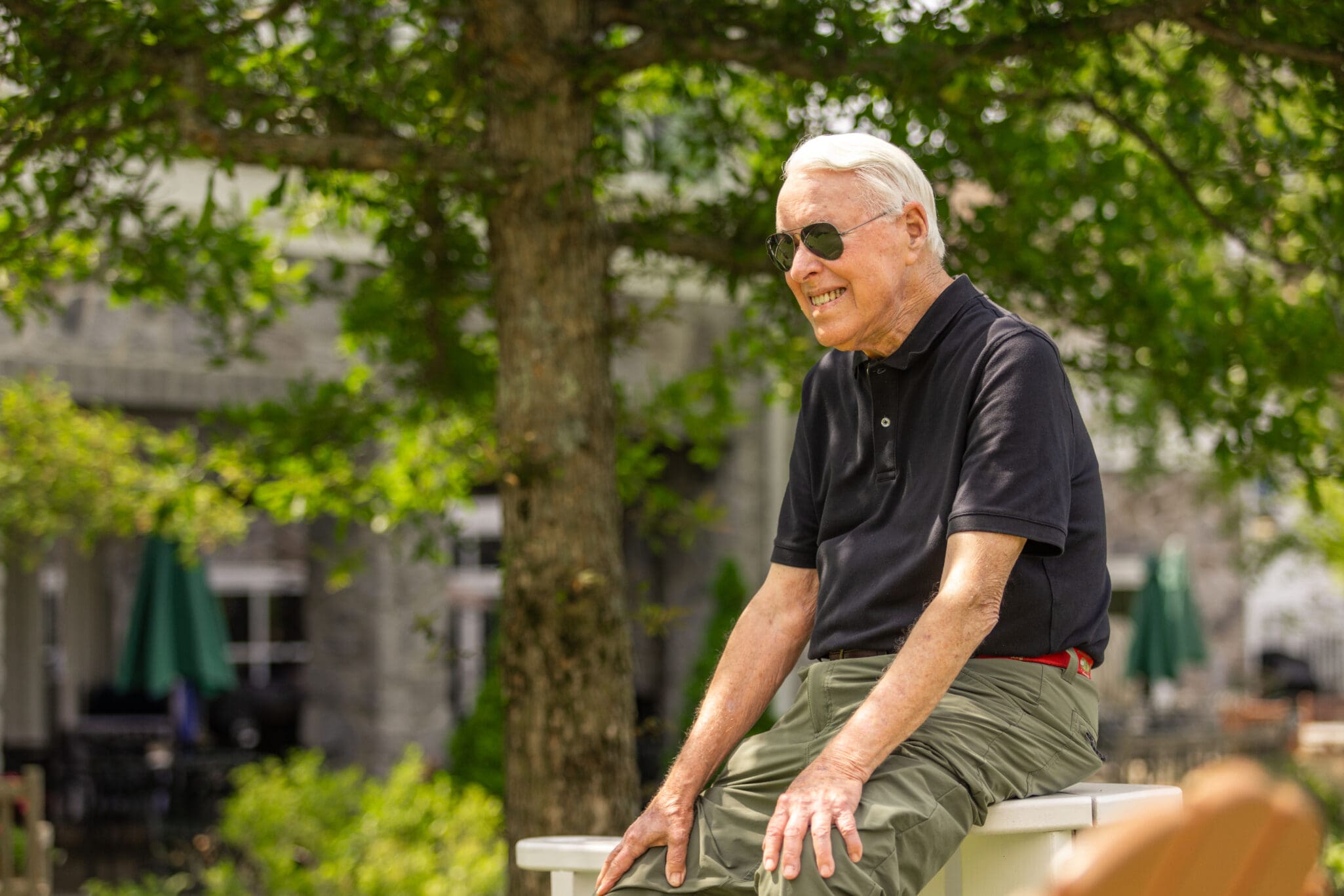 Senior man sitting outside and admiring growth at StoneRidge. The senior man is wearing a black shirt, sun glasses and green pants.