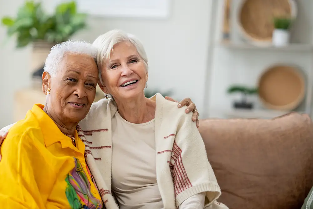 Two senior friends sit together on a sofa in their retirement residence. They are both dressed casually and huddled in closely as they embrace for the portrait.