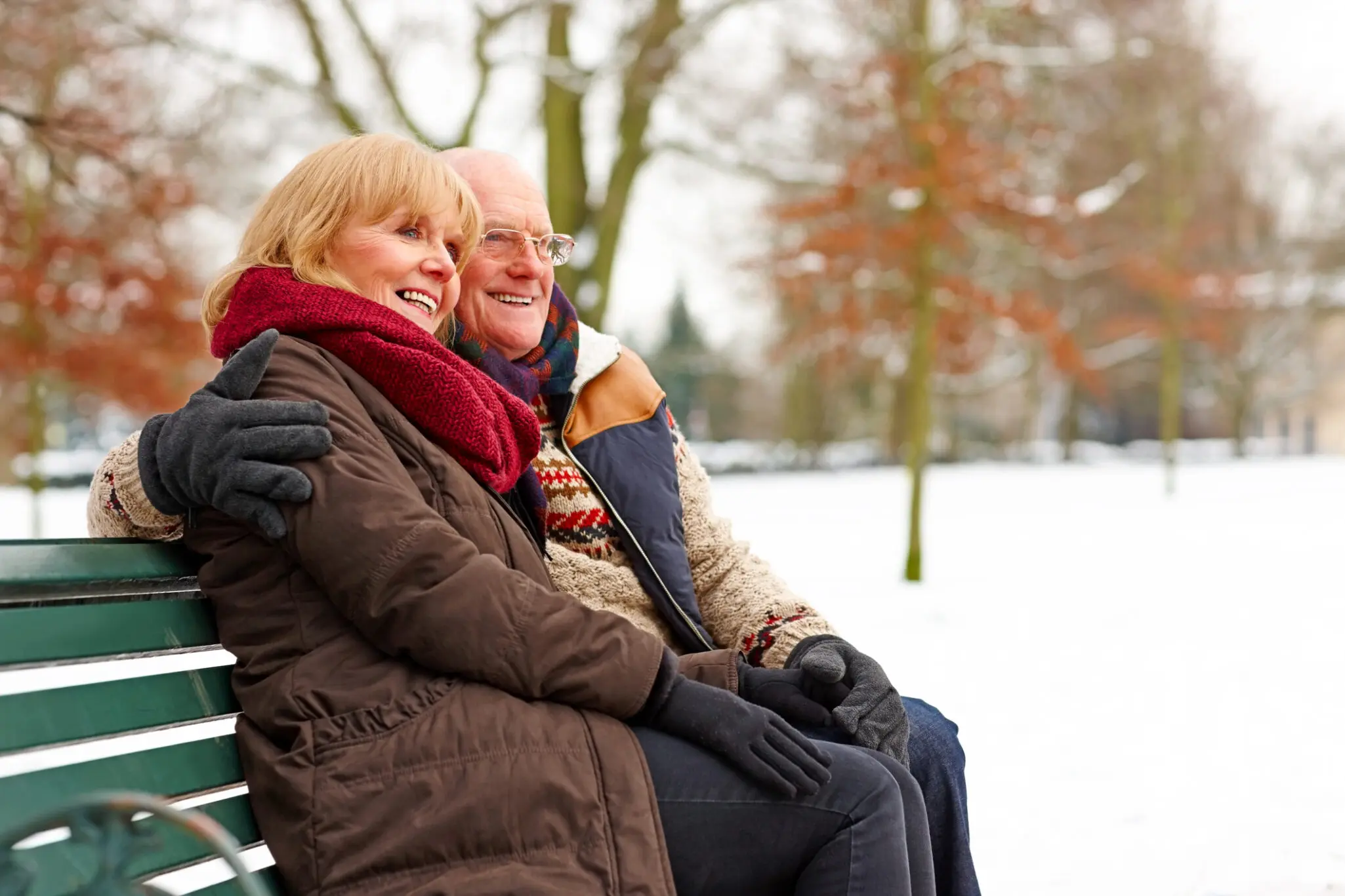 senior companionship: older couple wearing heavy jackets and scarves snuggles on bench in the snow